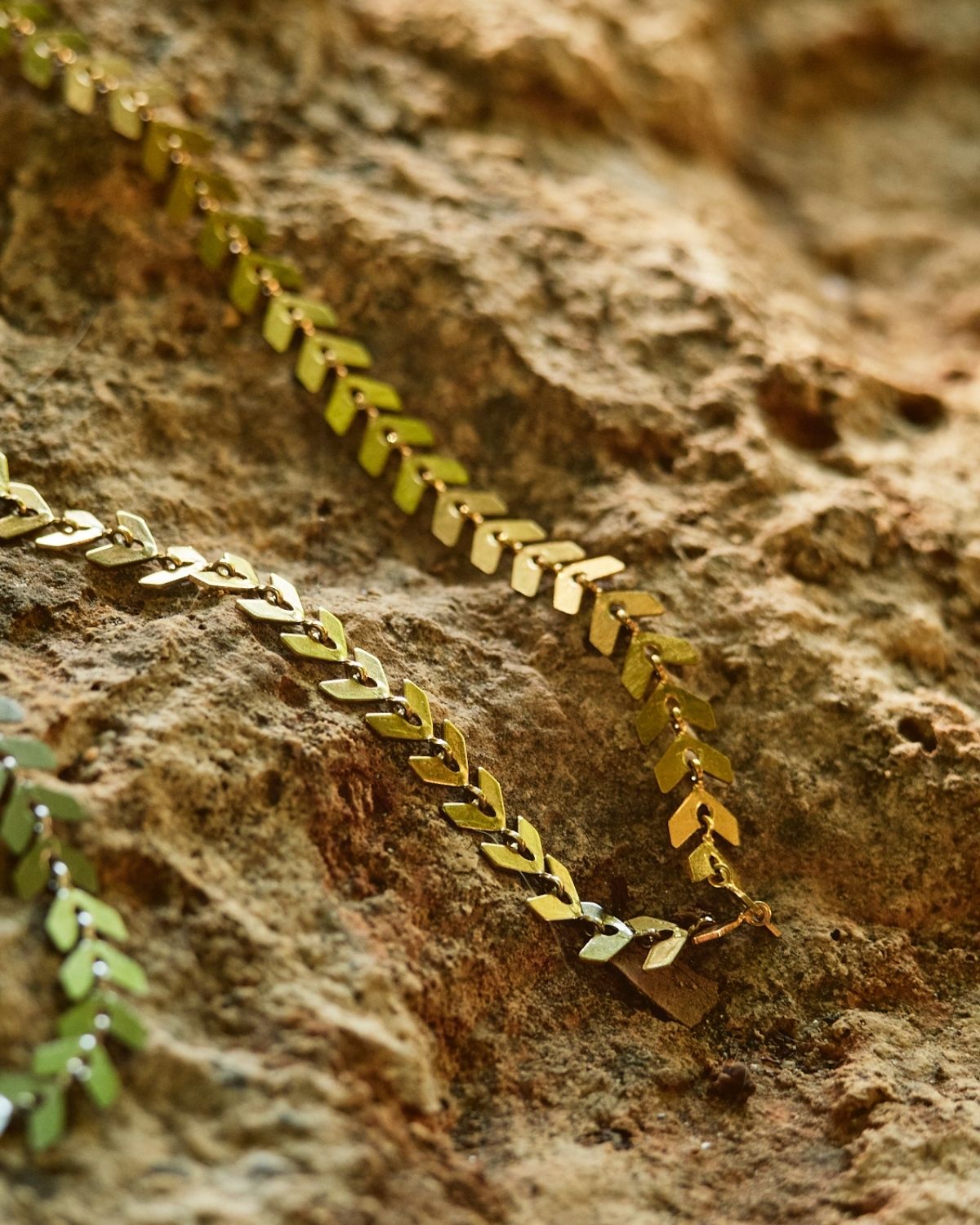 Stainless steel arrow necklace – Gold Close-up of a gold leaf design necklace placed on a rocky surface